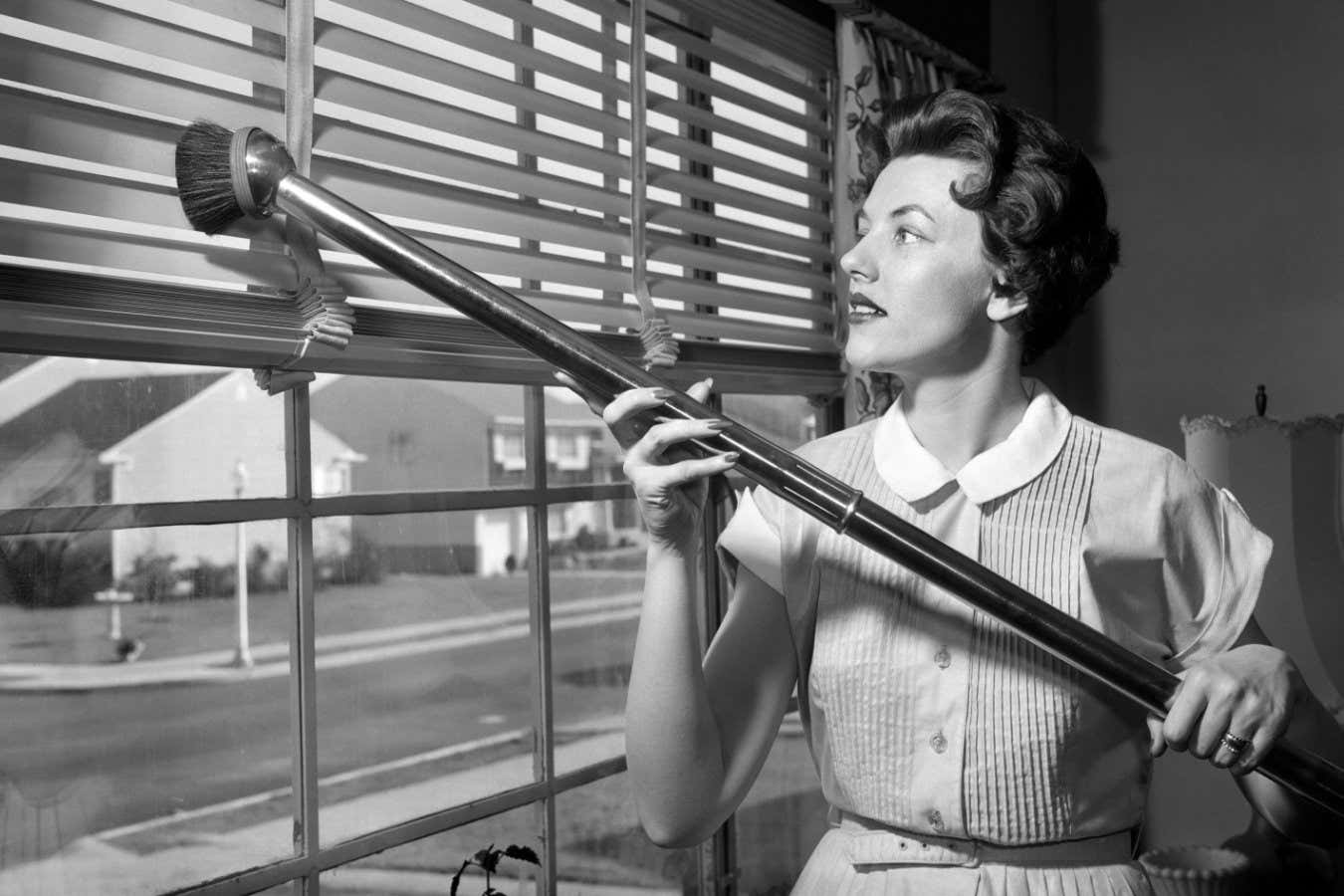F14FTY 1950s WOMAN VACUUMING VENETIAN BLINDS ON WINDOW LOOKING OUT ONTO SUBURBAN STREET