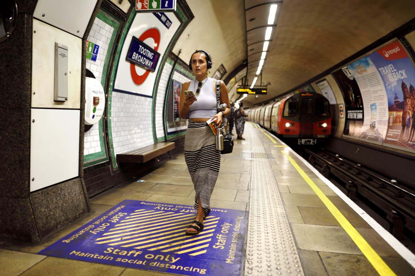 2JHRXY3 London, England, UK. London Underground - young woman with mobile phone and wearing headphone walking along the platform of Tooting Broadway station